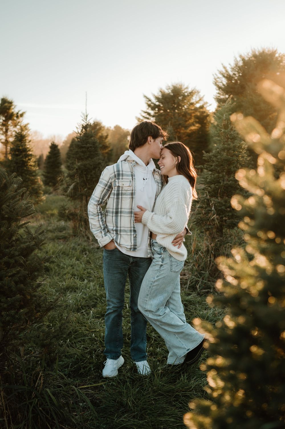 A romantic sunset moment between a couple standing among evergreen trees in casual fall attire.