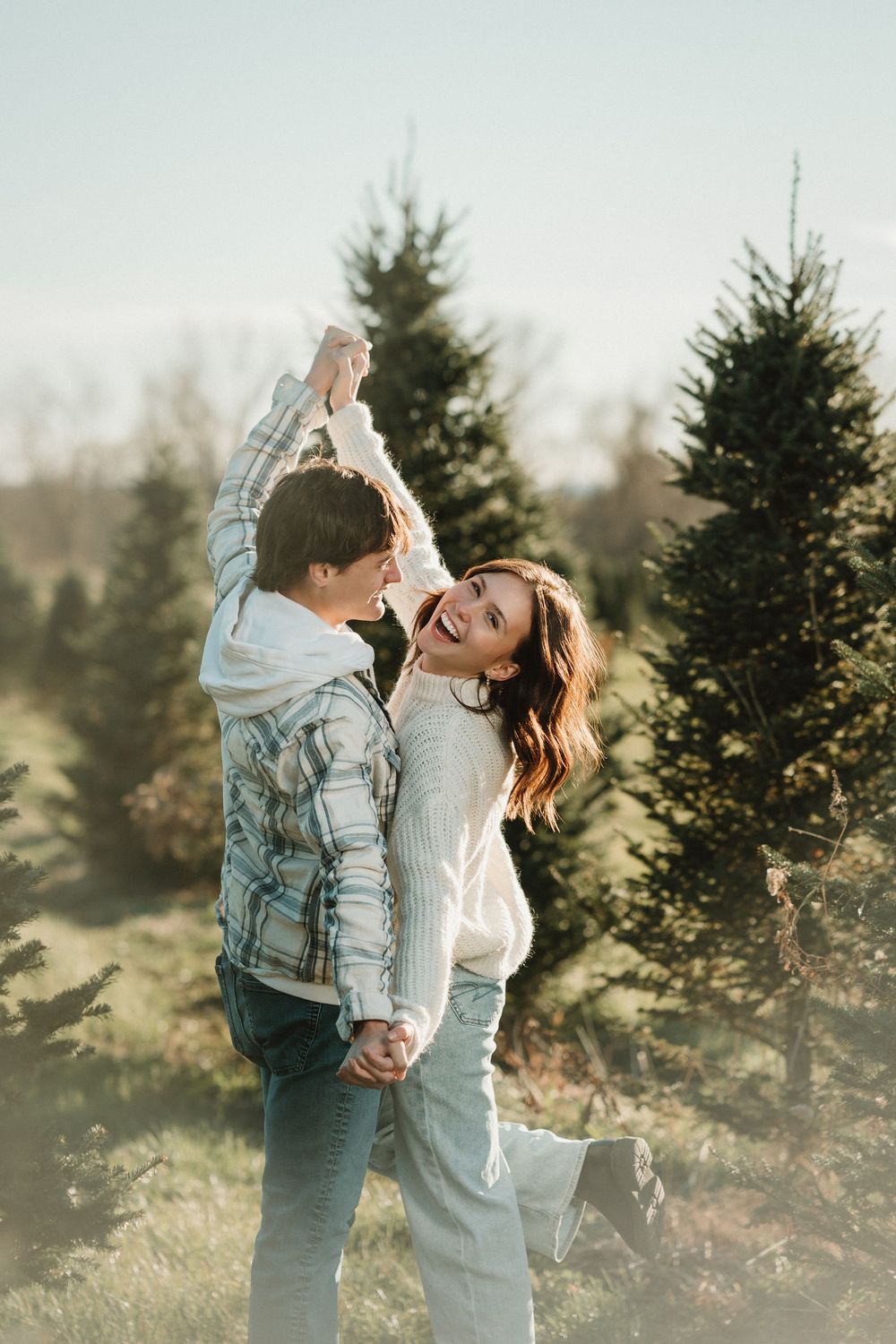 A joyful couple shares a romantic moment among evergreen trees at sunset, dancing and embracing in warm light.