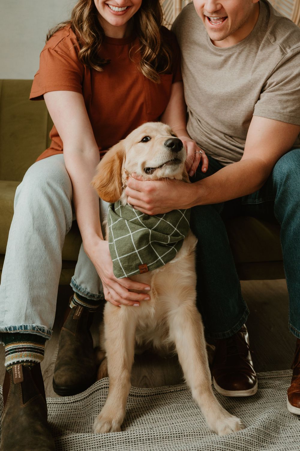 A couple sits on a couch with their golden retriever puppy wearing a green sweater in a warm indoor setting.