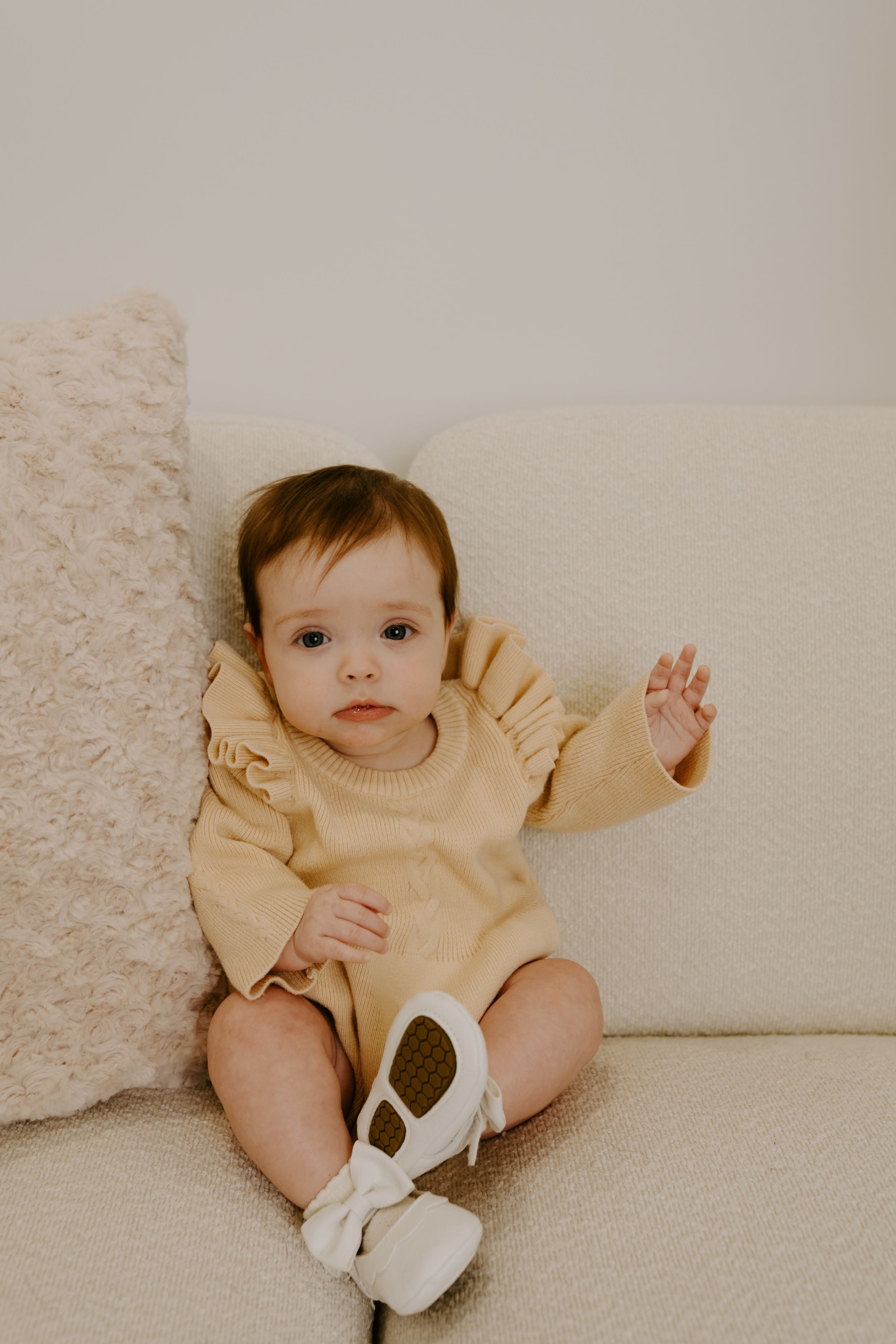 A baby wearing a cream-colored outfit sits on a white textured surface.
