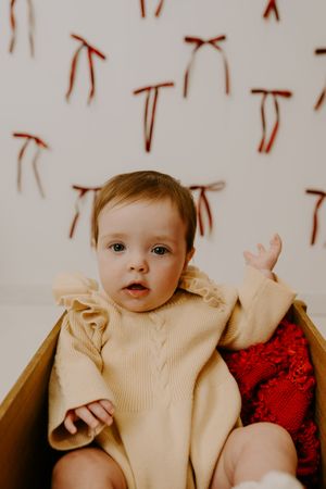 Baby wearing cream colored outfit against white background with red bow accents.