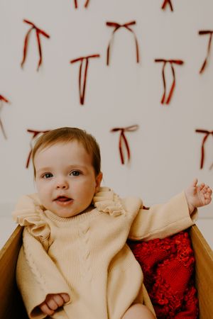 Adorable baby in cream outfit posing against white background with red decorative elements.