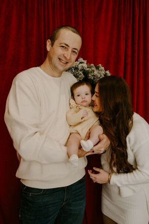 Candid family moments captured against red velvet curtain with baby's first photo shoot.