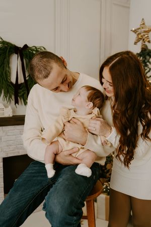 A tender family moment shared between parents and baby in front of a Christmas tree and fireplace.