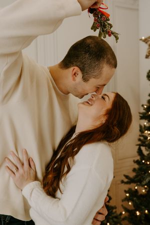 A romantic couple shares an intimate moment under mistletoe with Christmas decorations in the background.