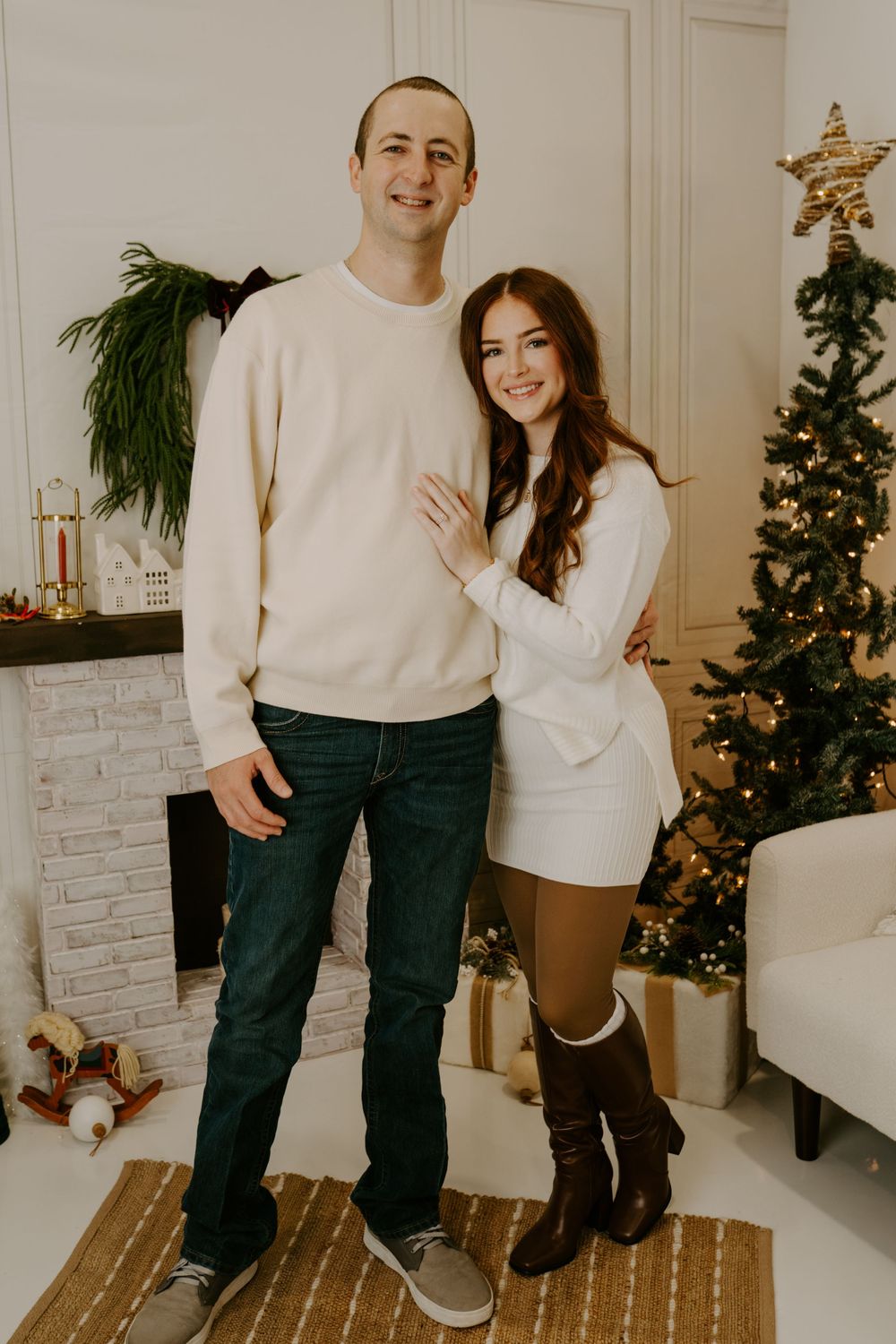 A couple in matching cream sweaters poses in front of their decorated Christmas tree and fireplace.
