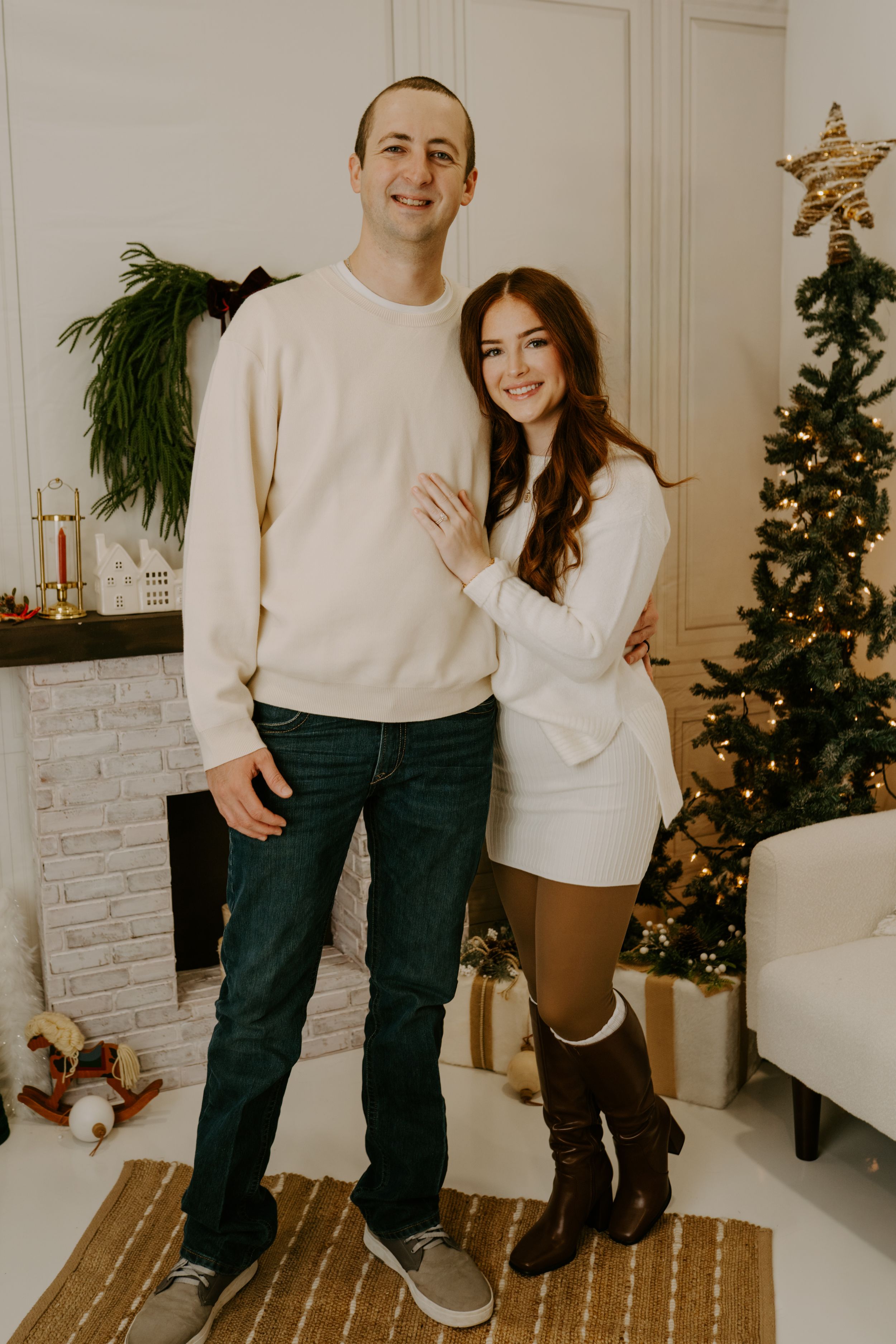 A couple in matching cream sweaters poses in front of their decorated Christmas tree and fireplace.