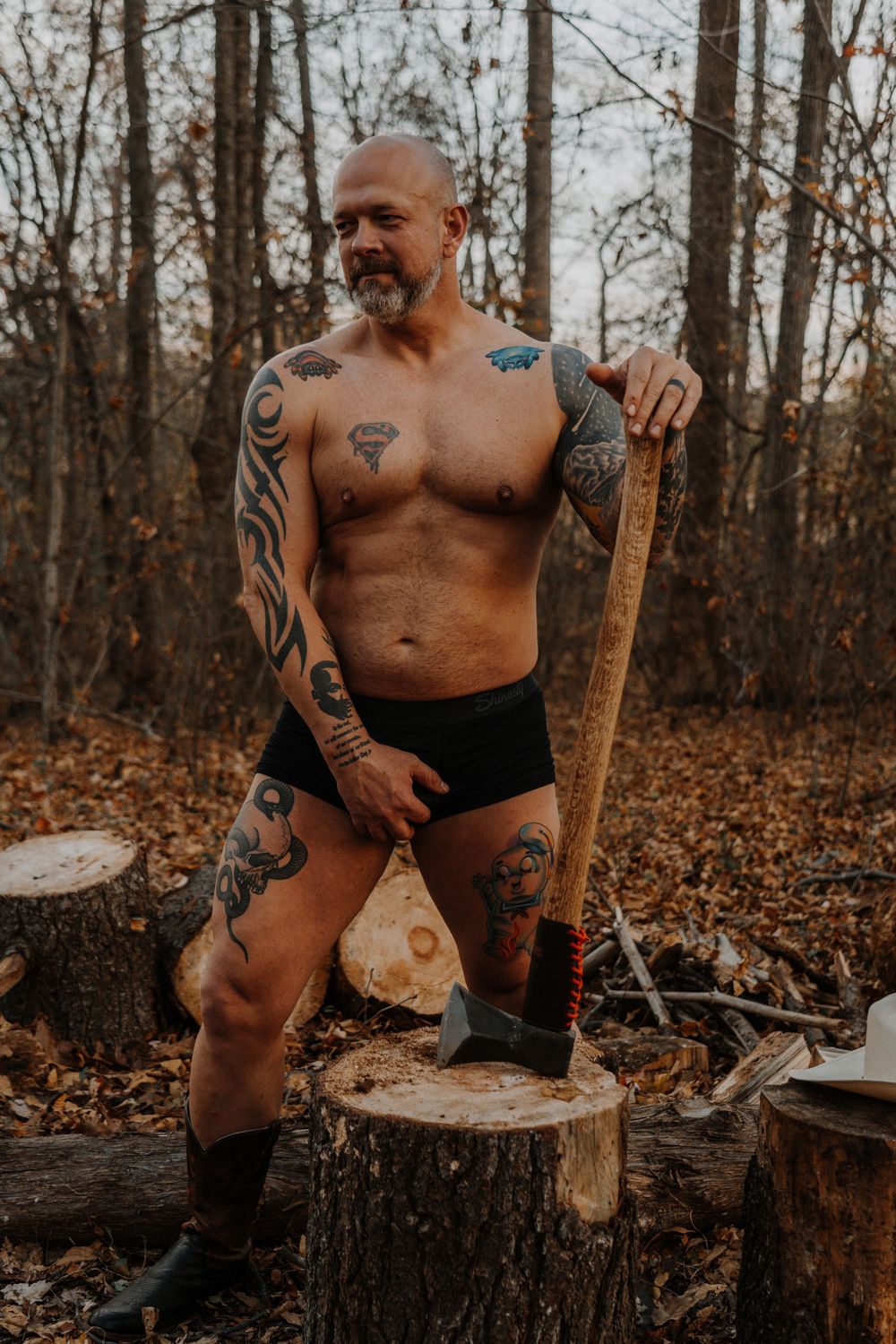 Shirtless person with tattoos sits on rustic wooden stump in autumn forest setting with fallen leaves.
