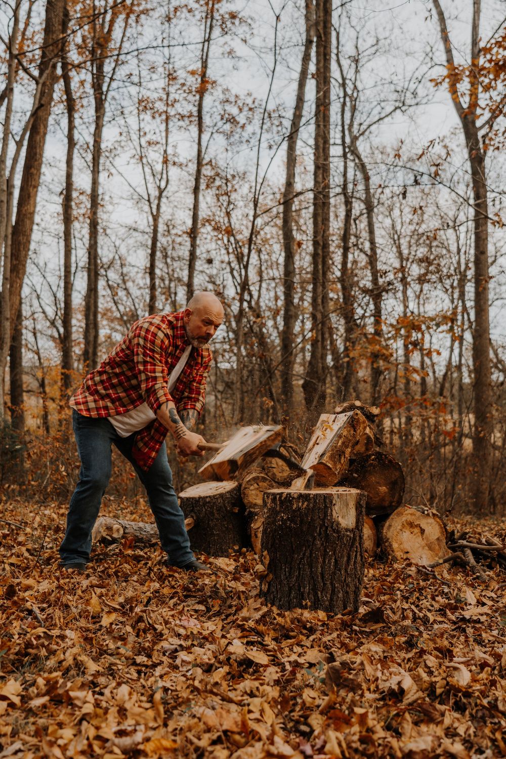 Someone in a plaid shirt chops wood in an autumn forest surrounded by fallen orange leaves.