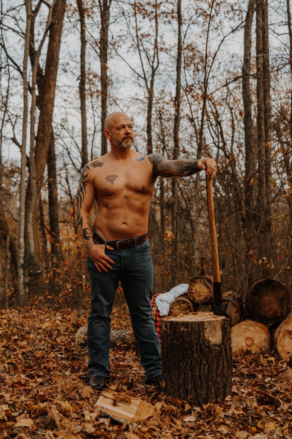 Shirtless man with tattoos stands in autumn forest next to fallen logs and autumn leaves.