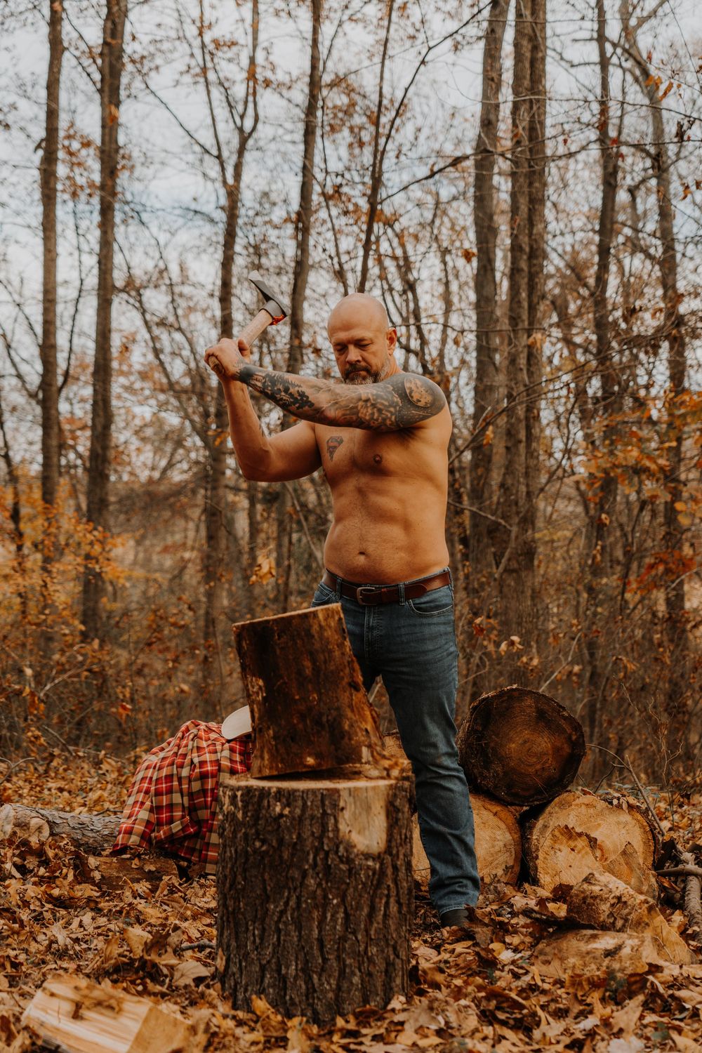 A shirtless man with tattoos chops wood in an autumn forest setting while wearing jeans.