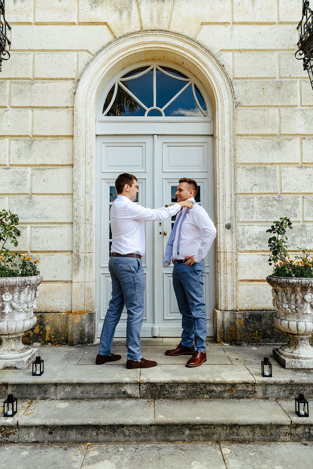 Two people in business casual attire stand laughing together on stone steps in front of an elegant white doorway.