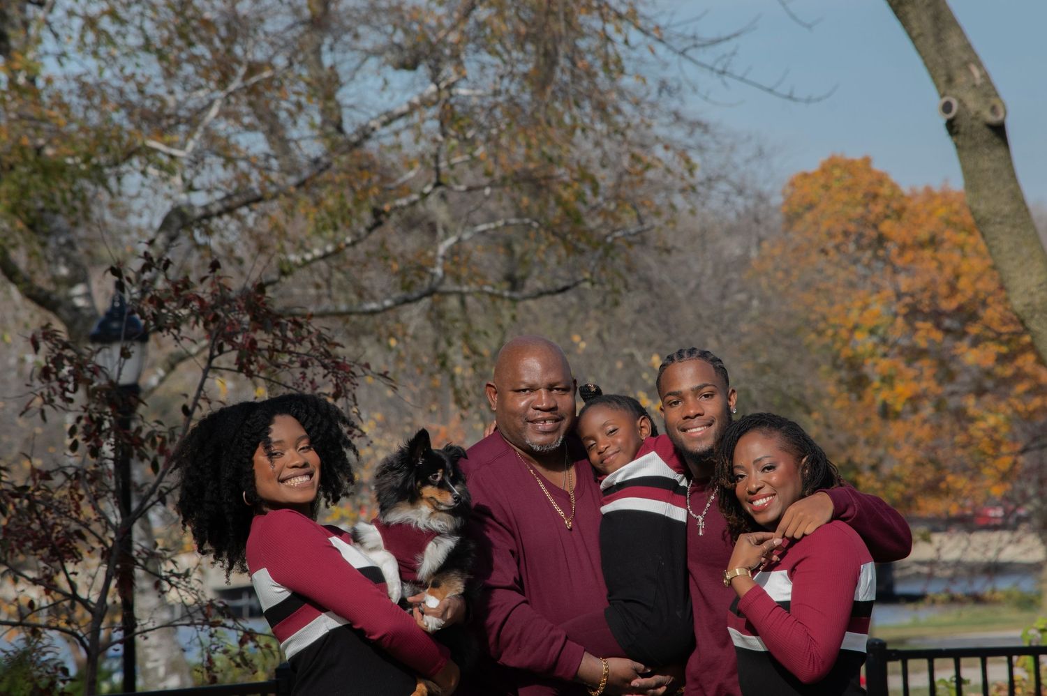 A family wearing matching burgundy sweaters poses together outdoors during autumn with fall foliage in the background.