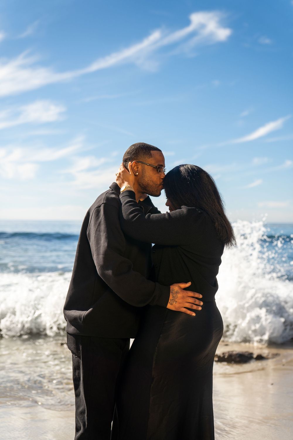 Two people embrace on a beach with crashing waves and blue sky in the background.