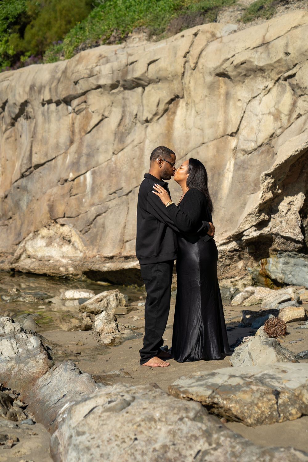 A couple shares a romantic moment on rocky coastal cliffs during a sunset photoshoot.