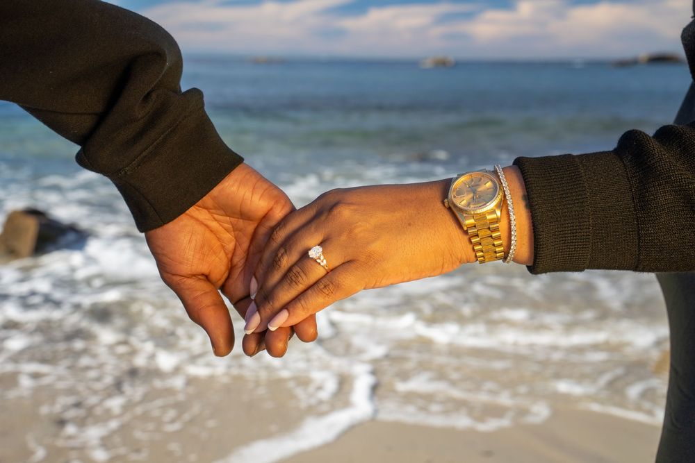 Two people holding hands on a beach with ocean waves and golden sunlight illuminating their clasped hands.