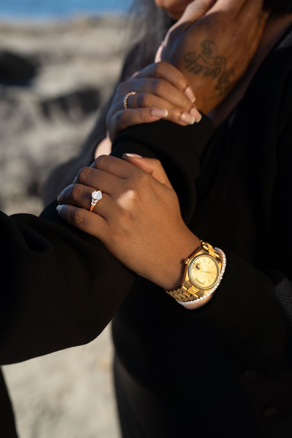 Close-up view of hands wearing engagement ring and gold watch with blurred background.