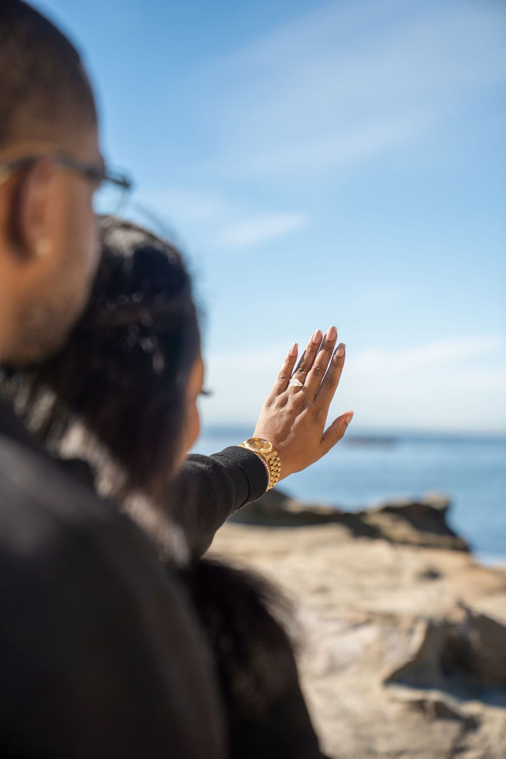 A raised hand with a gold bracelet reaches toward the blue sky at a rocky beach with ocean views in the background.