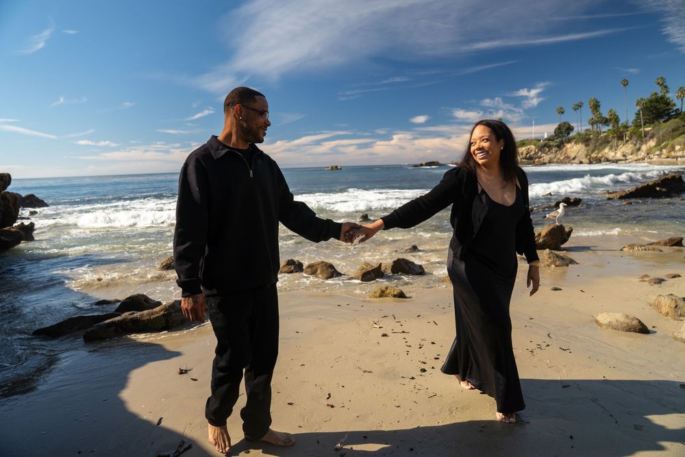 A couple walks hand in hand along a scenic rocky beach under blue skies and white clouds.