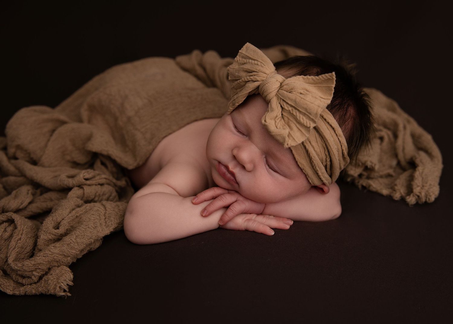 Peaceful sleeping newborn wrapped in soft brown fabric with matching bow headband on dark background.