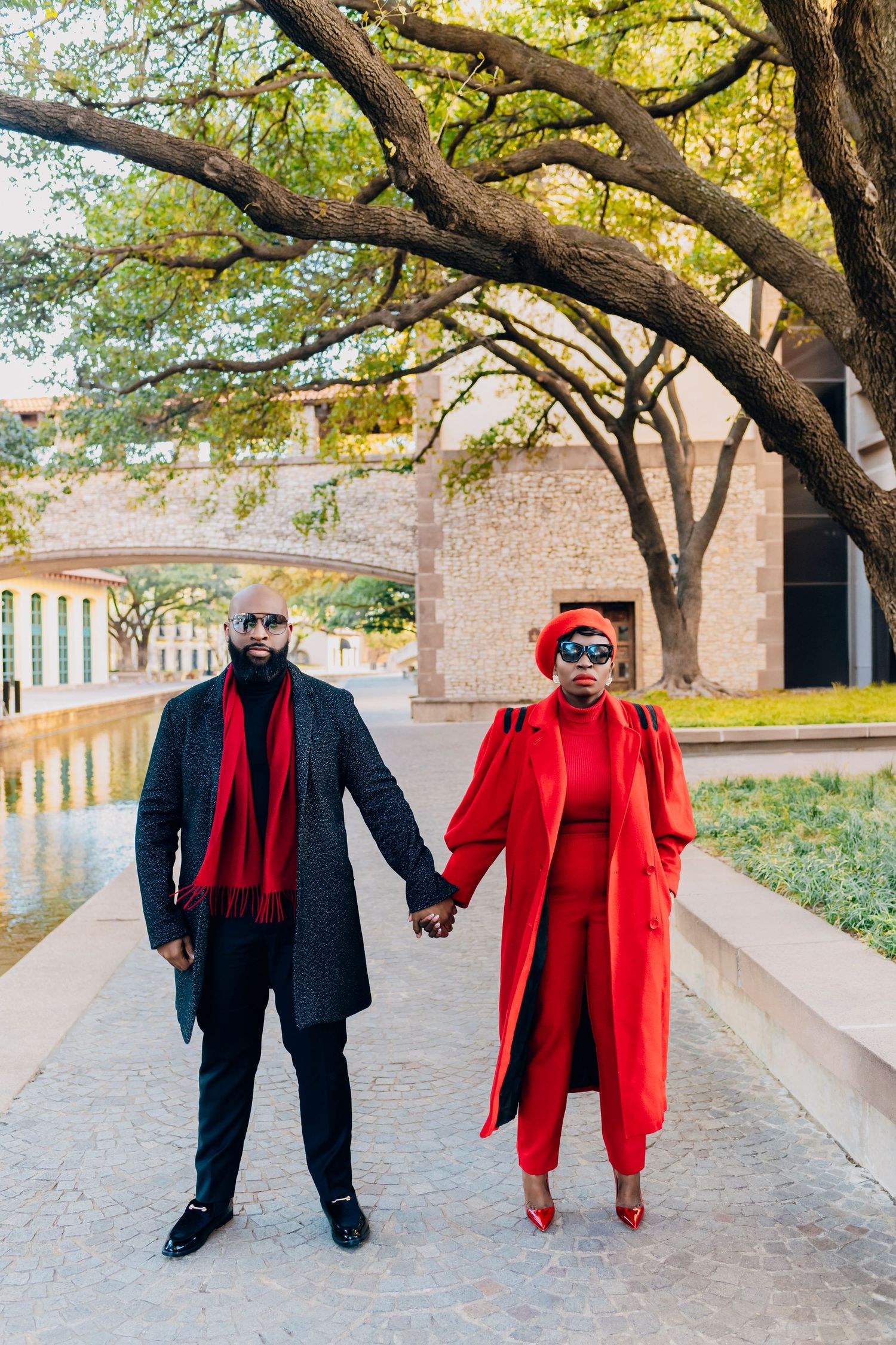 Two people in coordinated winter outfits holding hands on a walkway with red and navy coats, surrounded by fall trees.