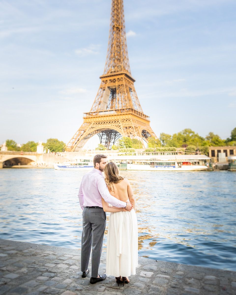 A couple stands together by the Seine River in Paris with the Eiffel Tower glowing warmly in the background at sunset.