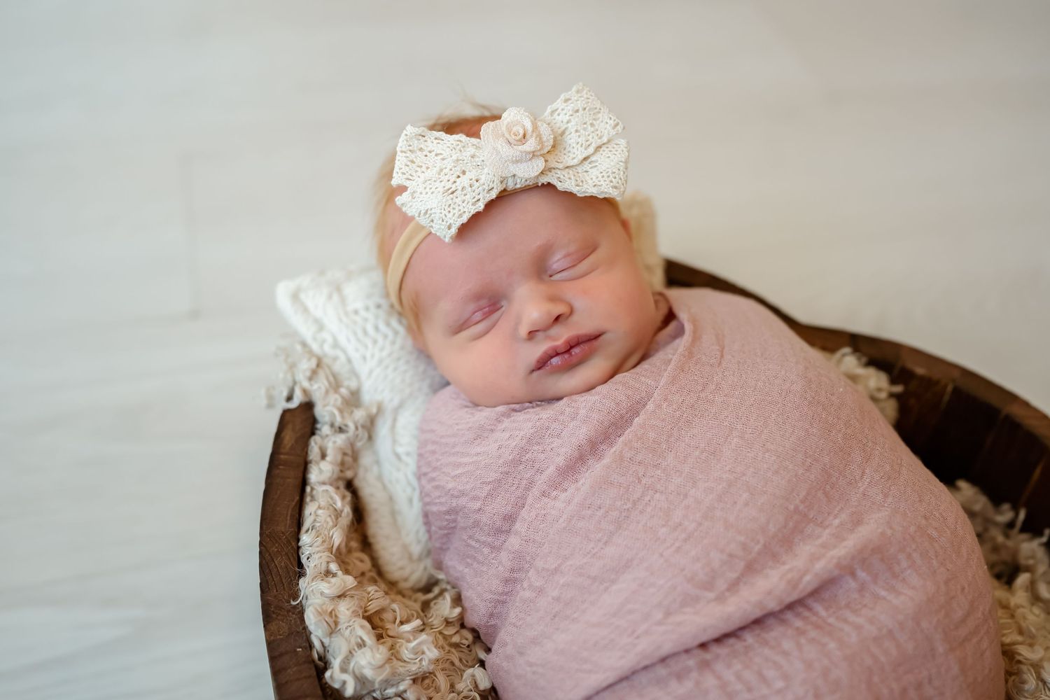Sleeping newborn baby wrapped in pink blanket rests peacefully in wooden bowl with lace headband.