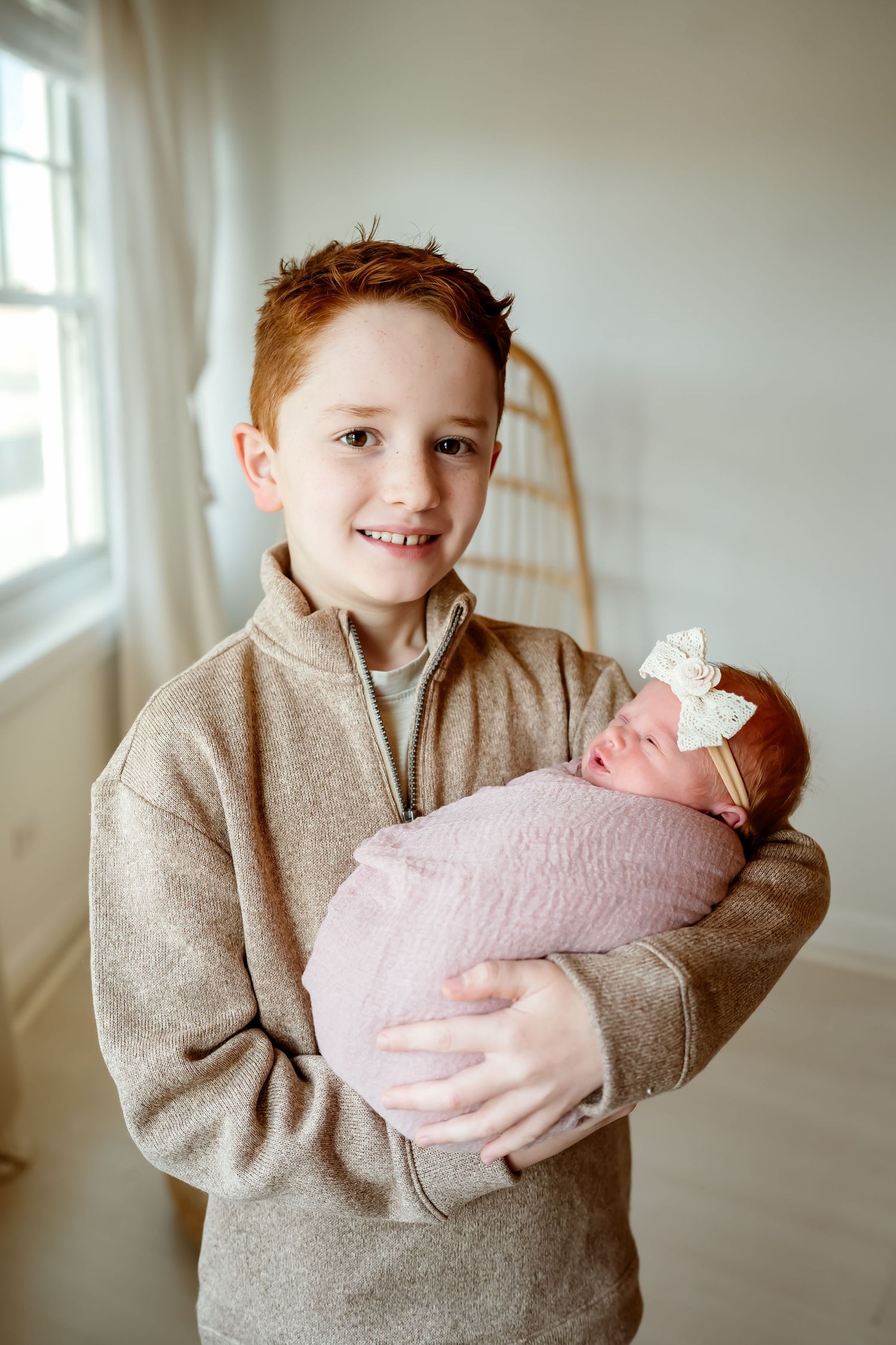A child in a beige sweater gently holds a newborn baby wrapped in soft pink blankets near a bright window.
