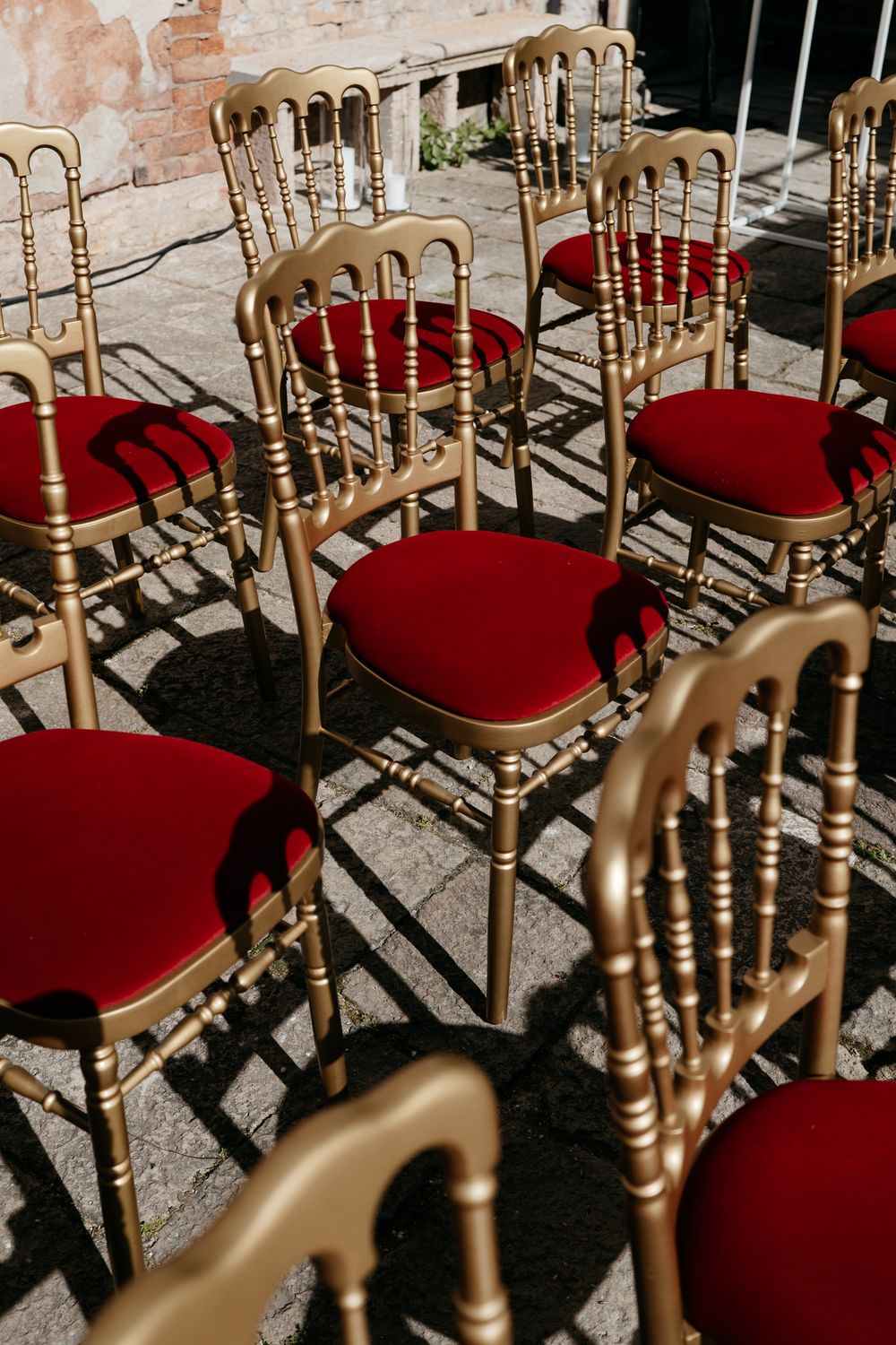 Gold Chiavari chairs with red velvet cushions arranged outdoors on stone pavement for an elegant event.
