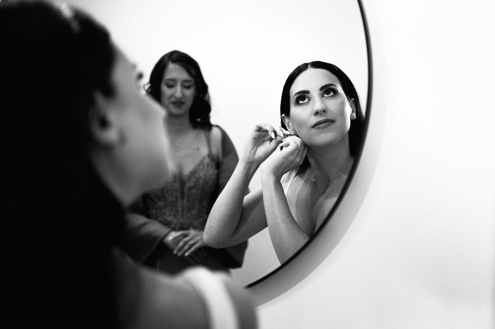 Black and white reflection in round mirror shows someone applying makeup while another person stands in background.