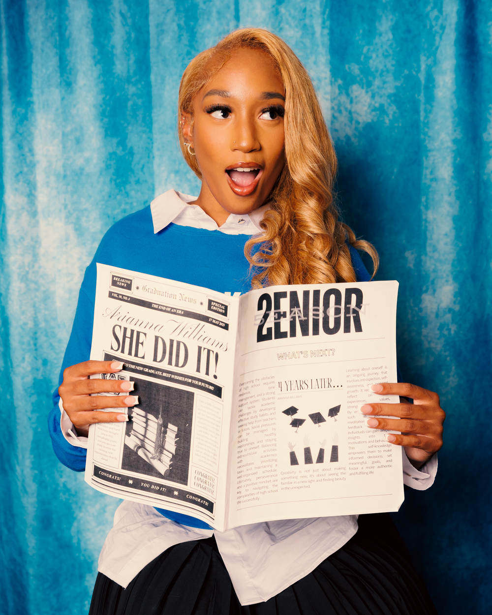 Student poses excitedly with open yearbook against turquoise background wearing white collared shirt.