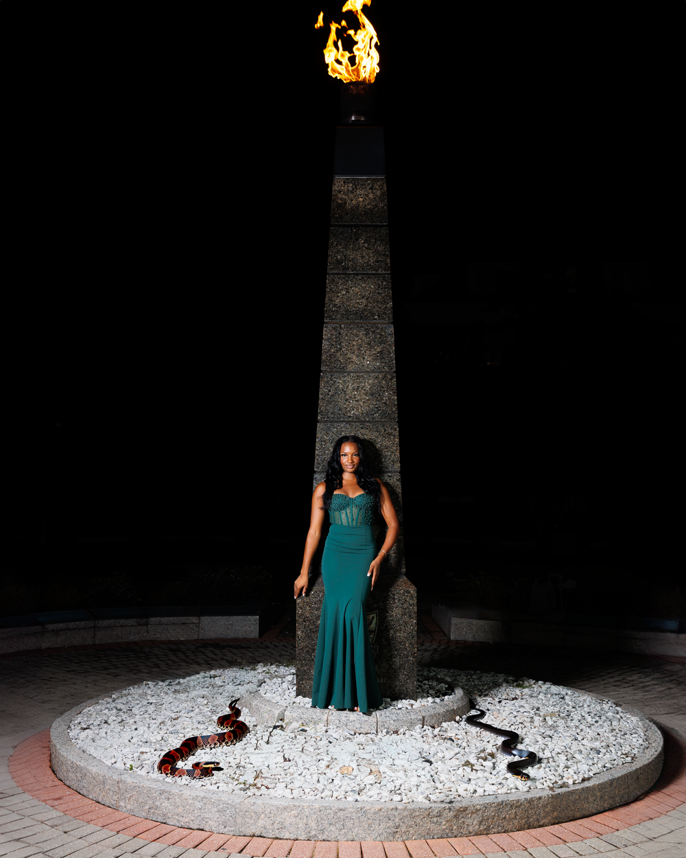 A woman in a teal evening gown poses against a dark backdrop with white stones and red accents on the ground.