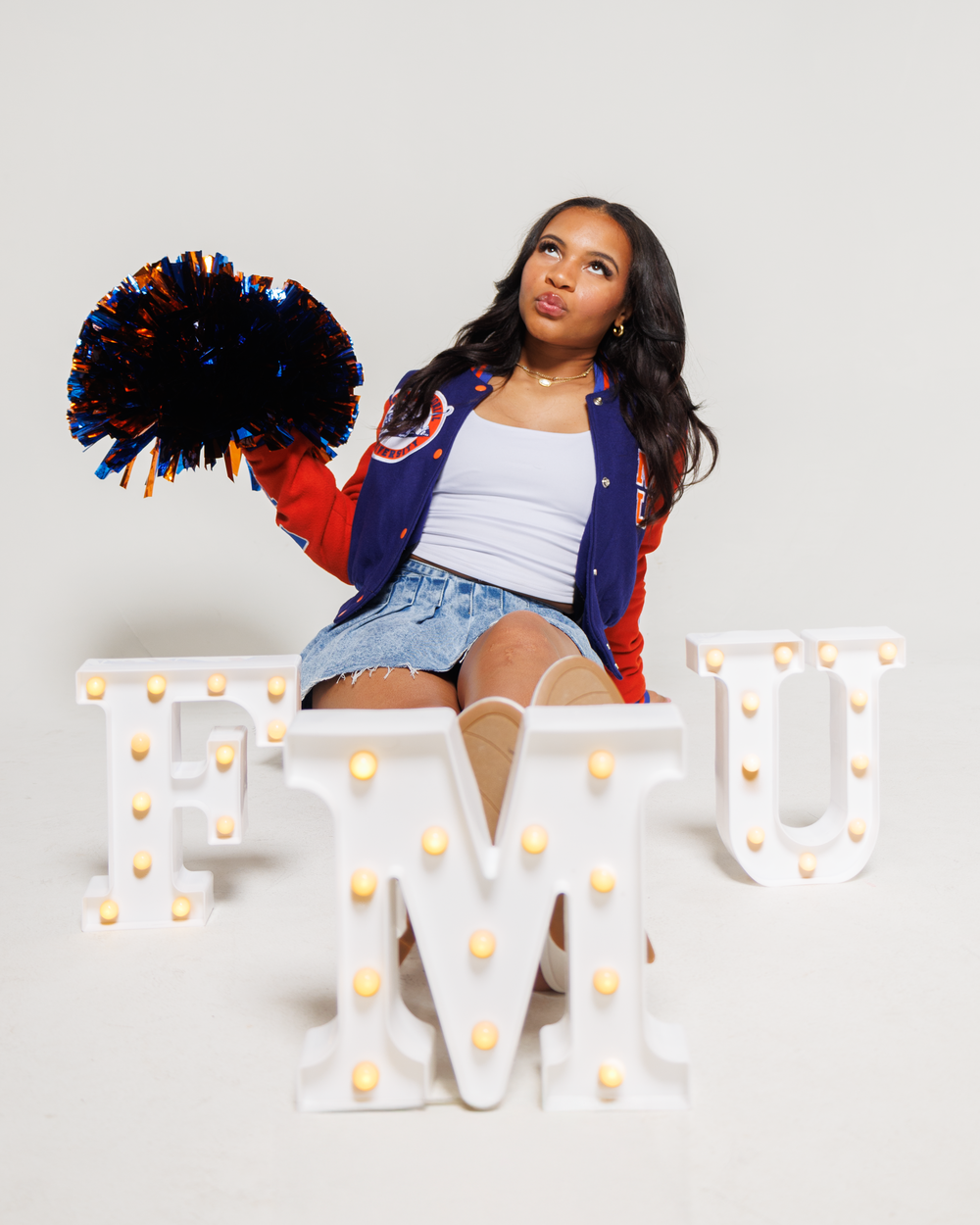 Person in white tank top and denim shorts poses with red and blue pom poms against white backdrop.