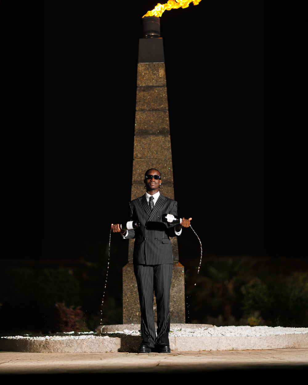 A figure in a dark suit stands dramatically lit against a black background with a white strip of light on the ground.