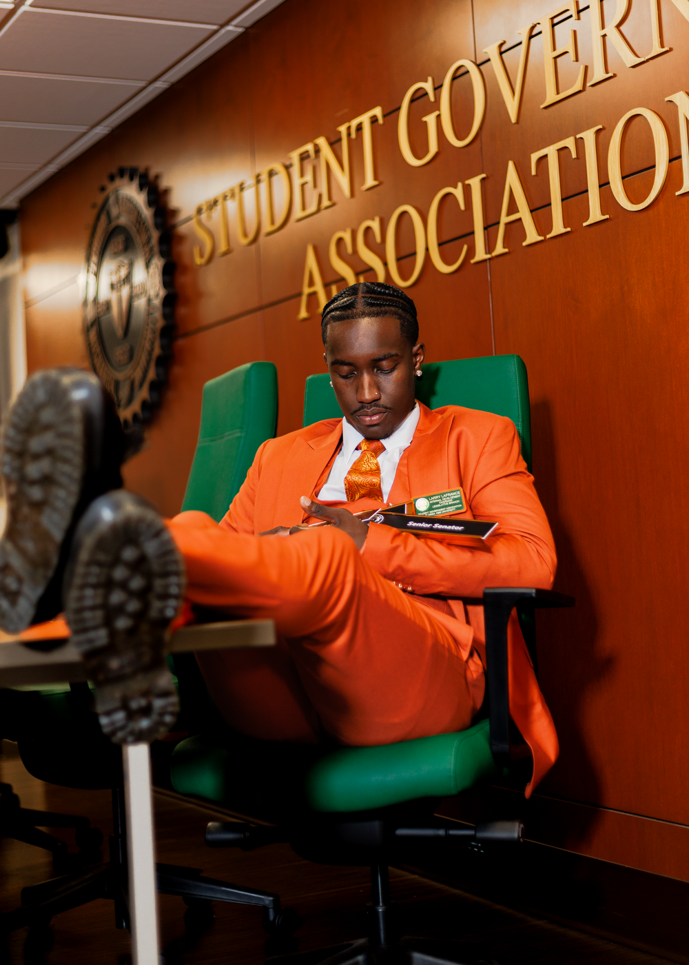 Student in orange blazer sits in green chair while studying and taking notes in university hallway.