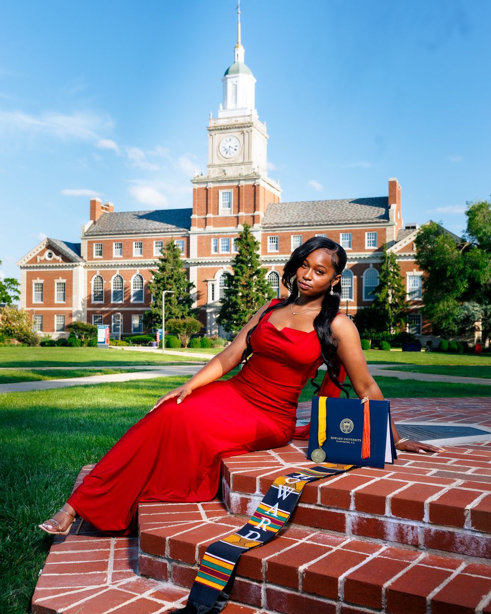 A graduate in a red dress poses with academic books on brick steps of a historic university building.