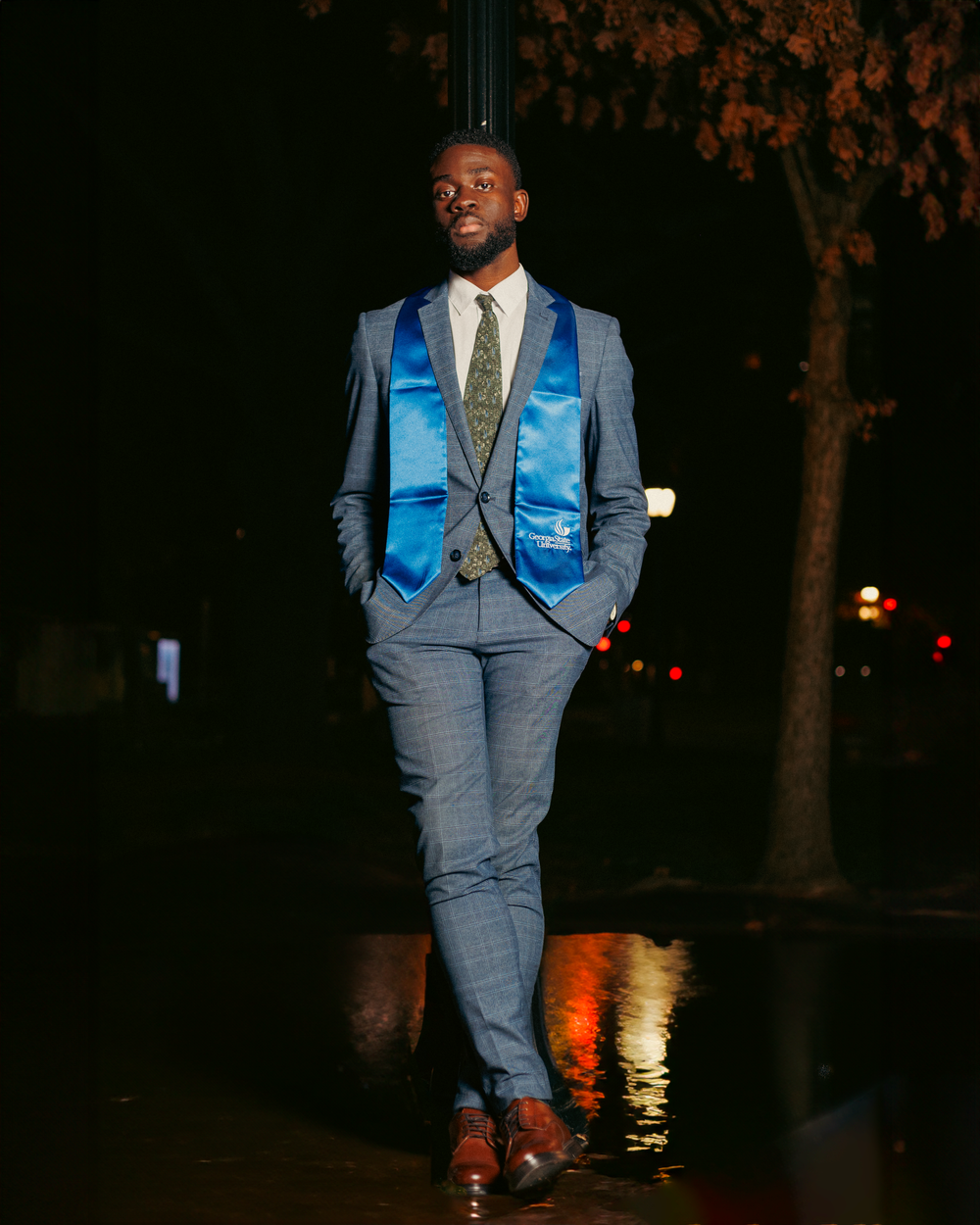 A person in a gray suit with light blue vest poses outdoors at night with illuminated trees in the background.