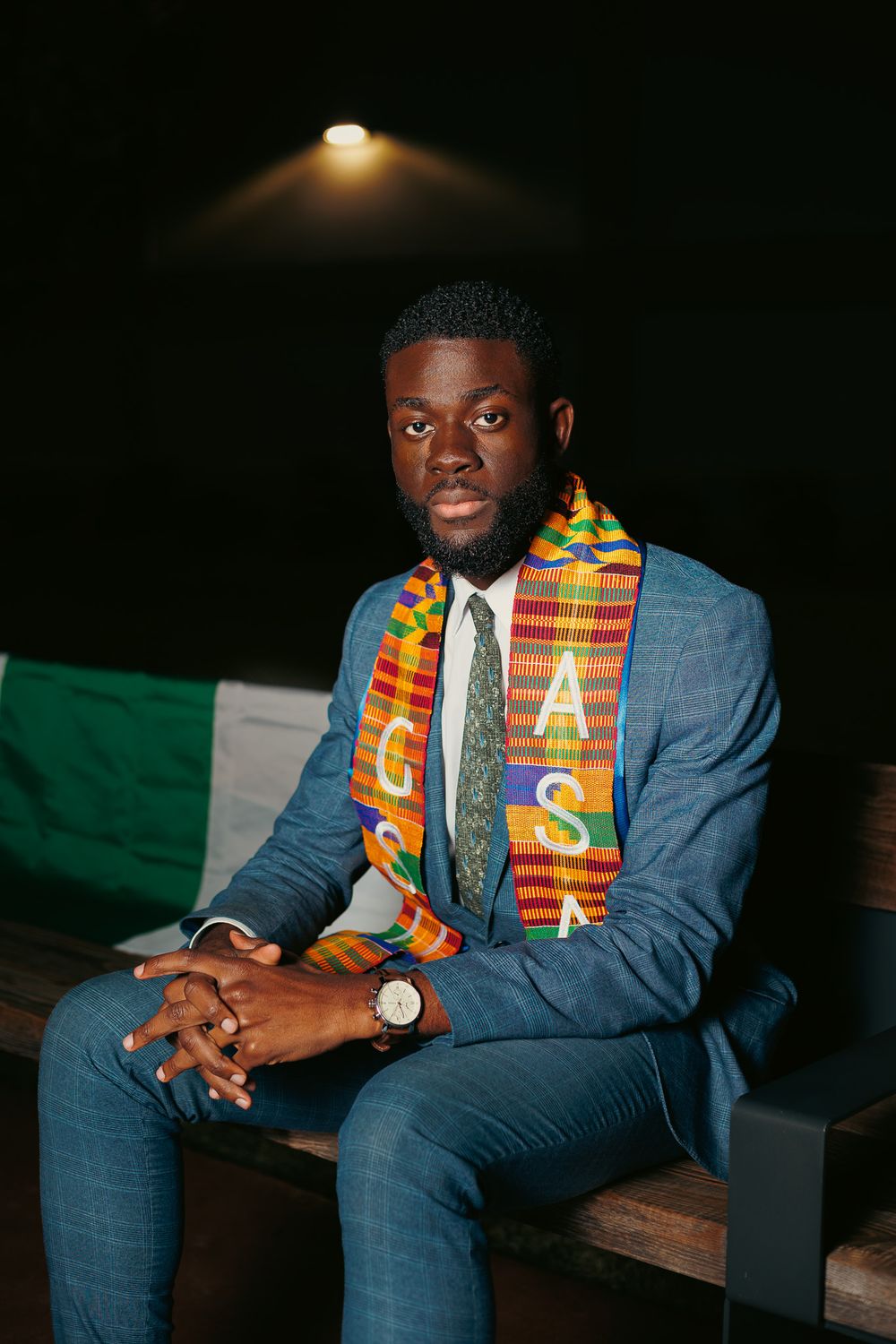 Person in formal attire with traditional kente scarf poses against dark background in studio setting.