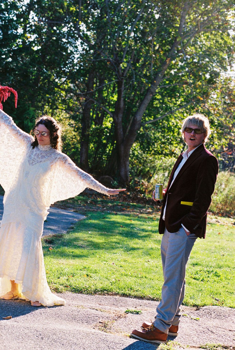 Bride holds out her arms in a full pose showing her wedding dress while the groom stands beside her looking into the camera, beer in hand.