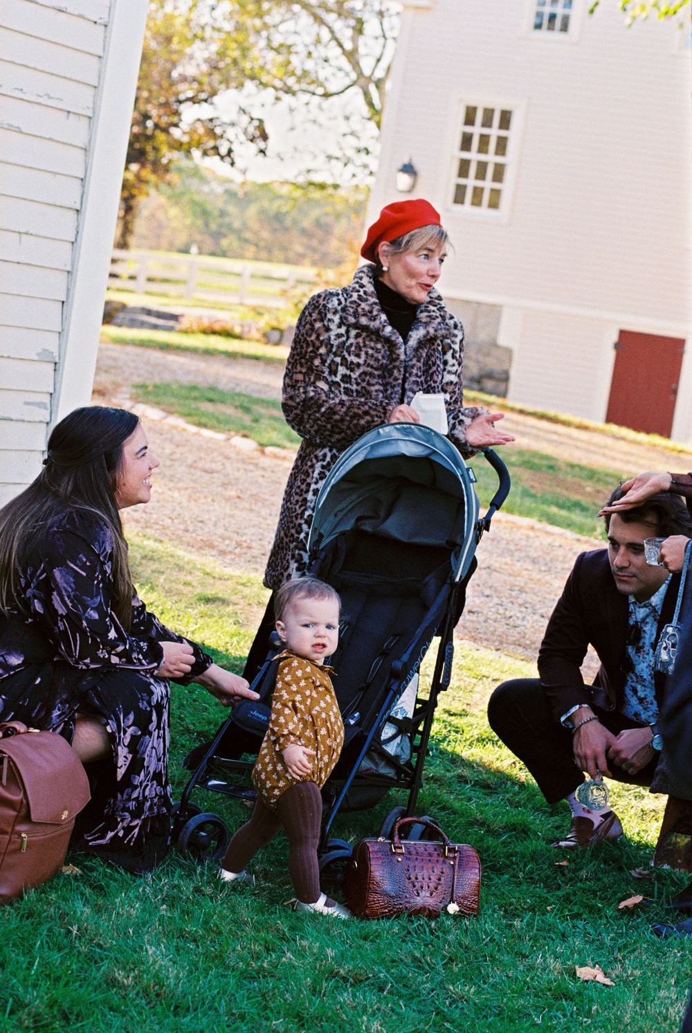 Woman in leopard coat and red beret stands with other guests and a stroller while the toddler stares at the camera outside after a wedding.
