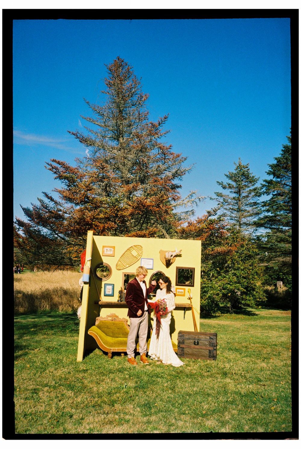 The couple poses outside in front of a vintage and colorful handmade Photo Booth with a couch and Wes Andersons style decorations in Maine.
