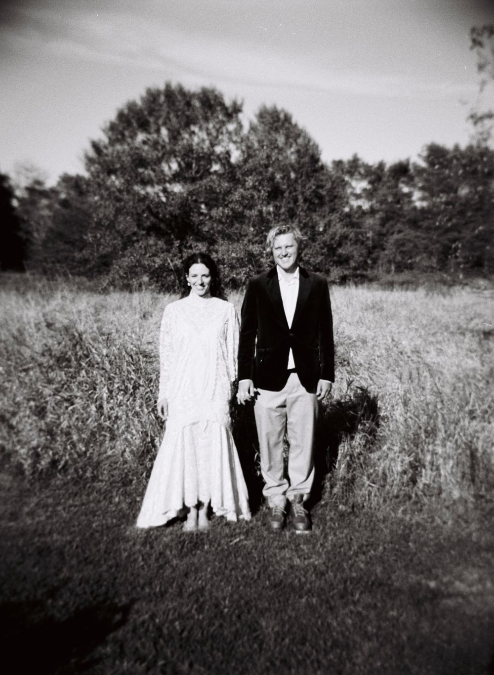 Just married couple stands together side by side facing the camera smiling in the sunshine in front of a field with trees in the background.