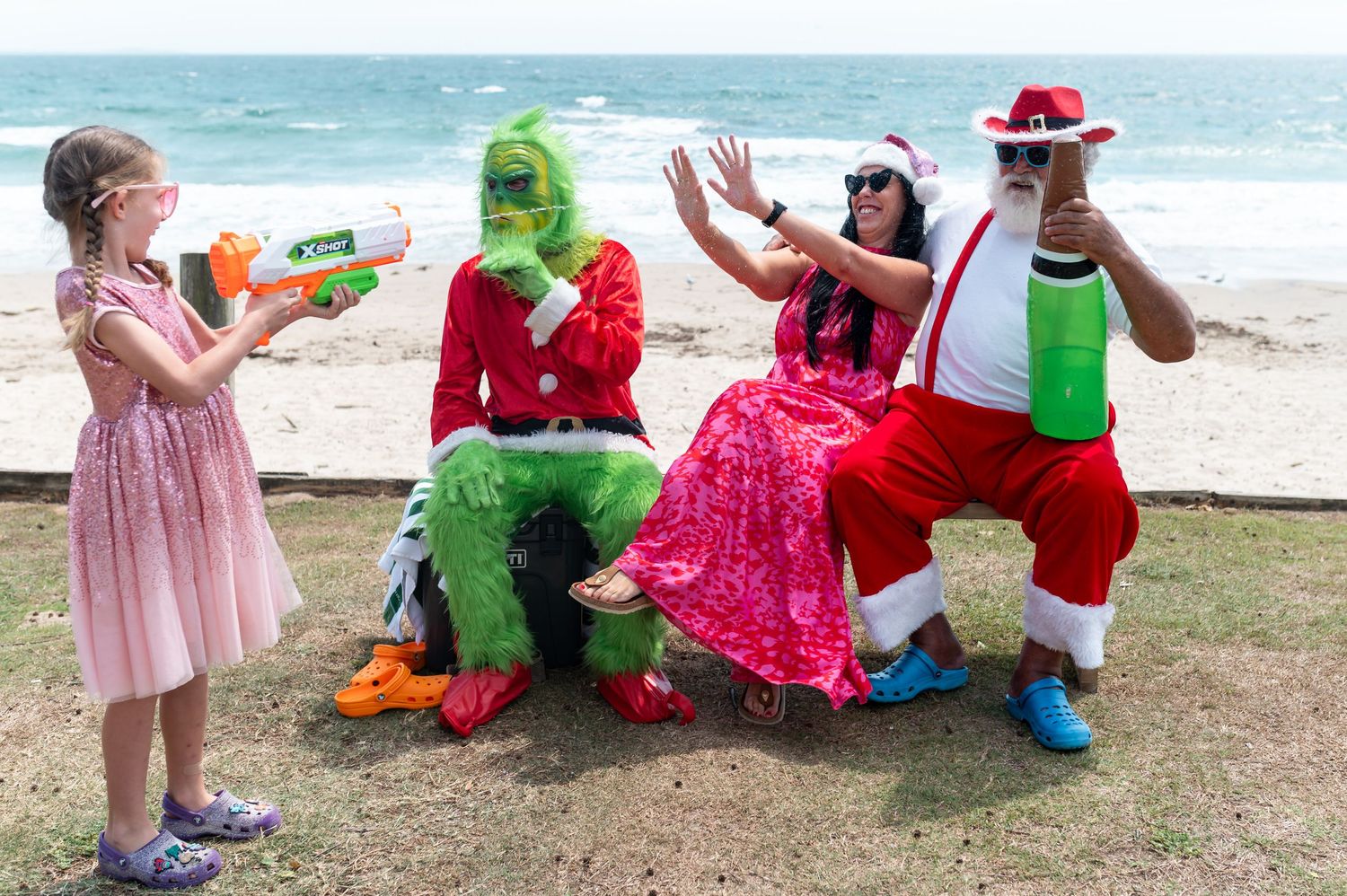 A group plays and interacts with a person in a green dinosaur costume on a grassy area near the beach.