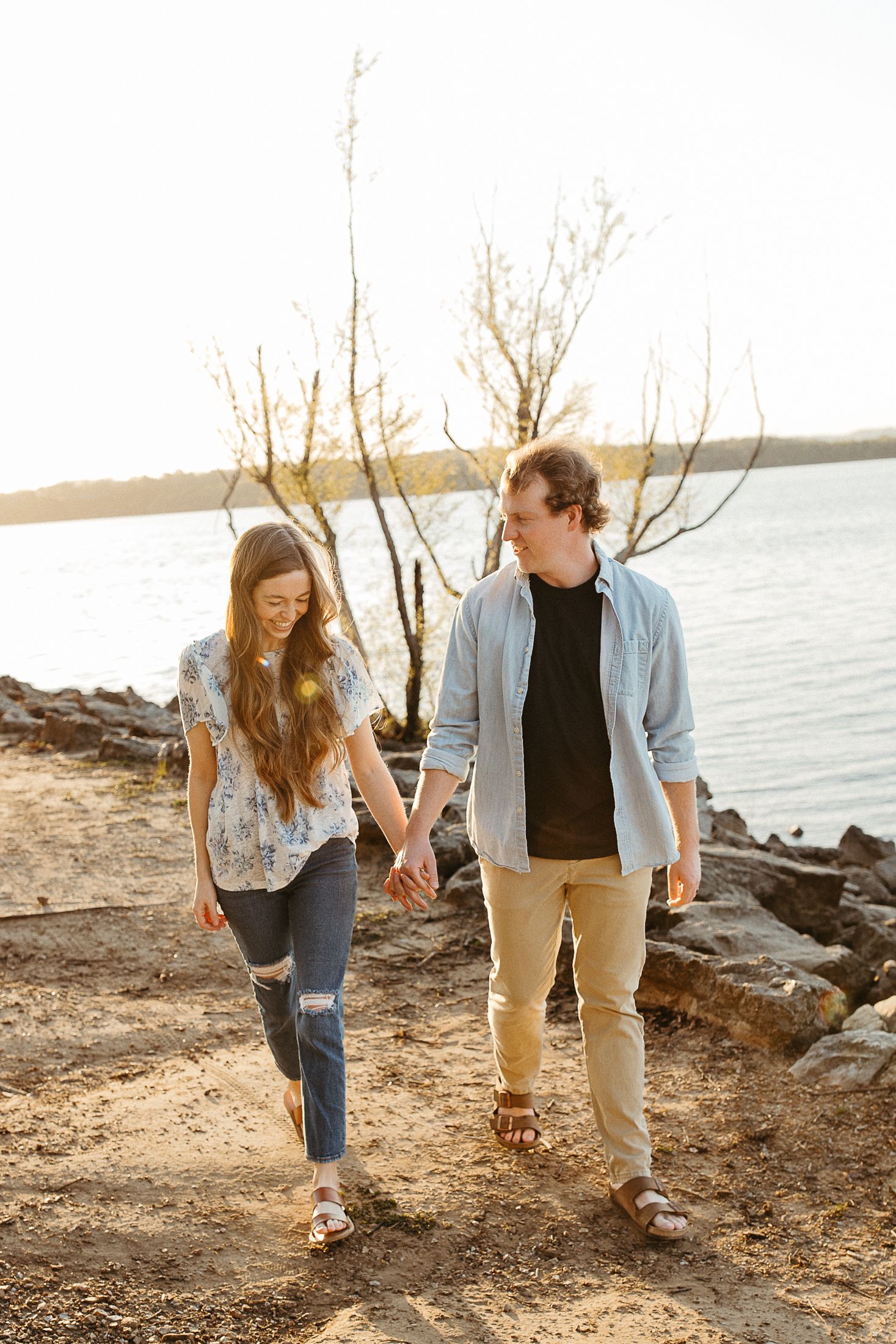 A couple walking hand in hand along a rocky beach shoreline at sunset in casual denim and dark clothing.