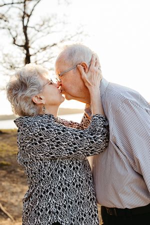 An elderly couple shares a tender kiss outdoors during golden hour, with sunlight filtering through trees in the background.