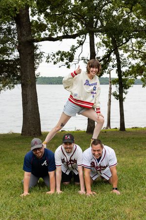 A group of people in baseball jerseys crawl on grass near a lake with trees in the background.