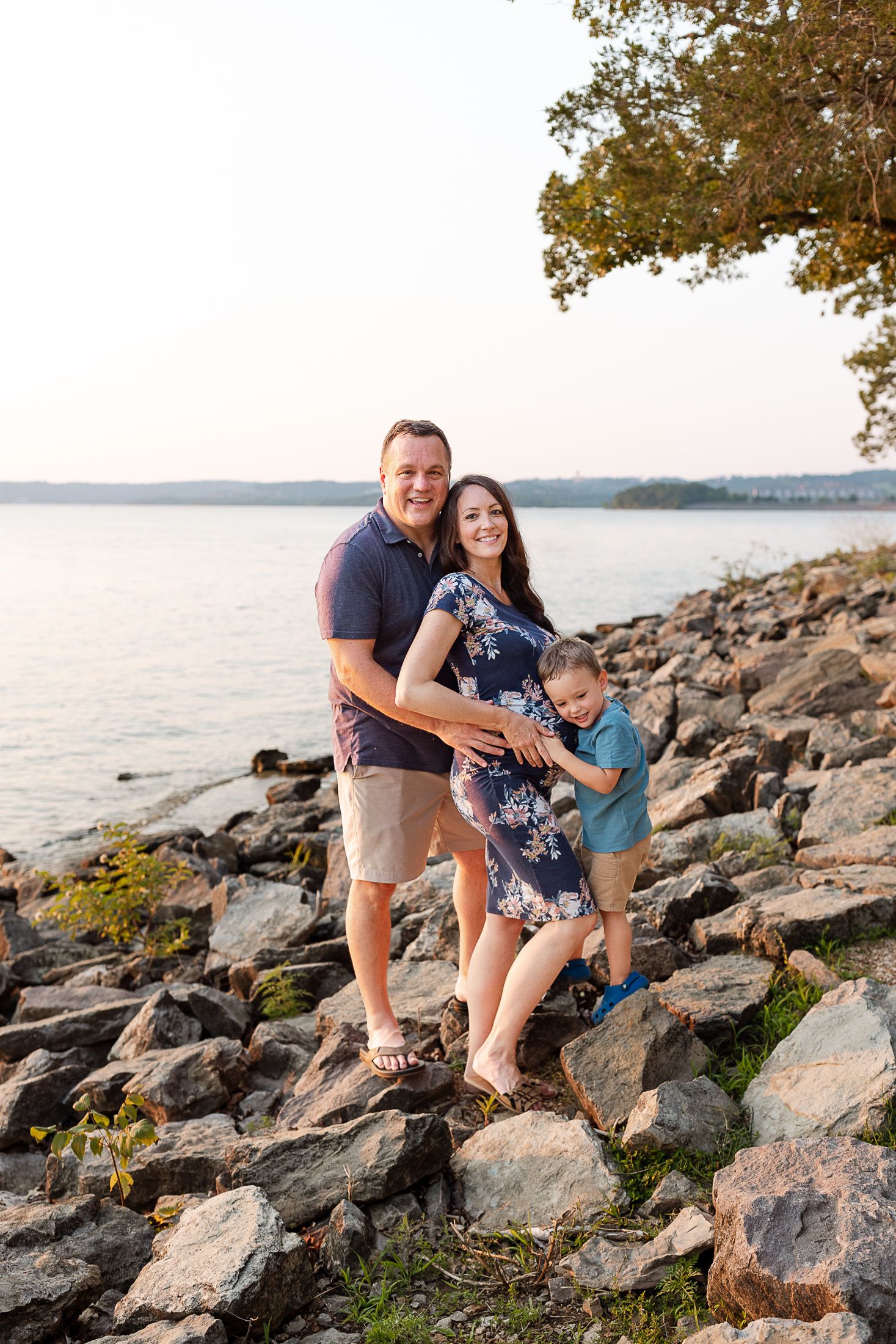 Family explores rocky shoreline together at sunset along peaceful waterfront with trees in background.