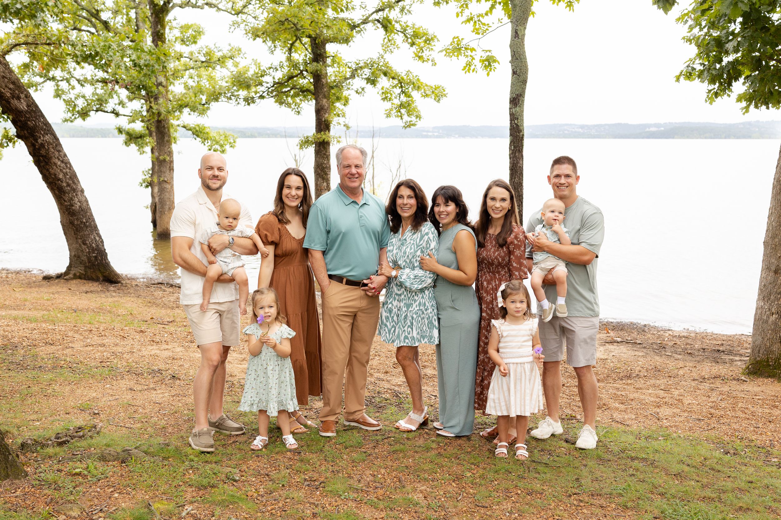 Extended family gathering outdoors near a lake with trees and natural scenery in the background.