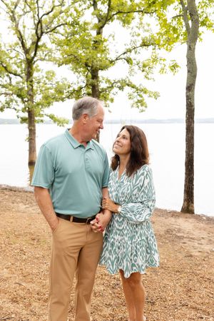 A couple shares a tender moment while walking along a lakeside path under green trees on a sunny day.