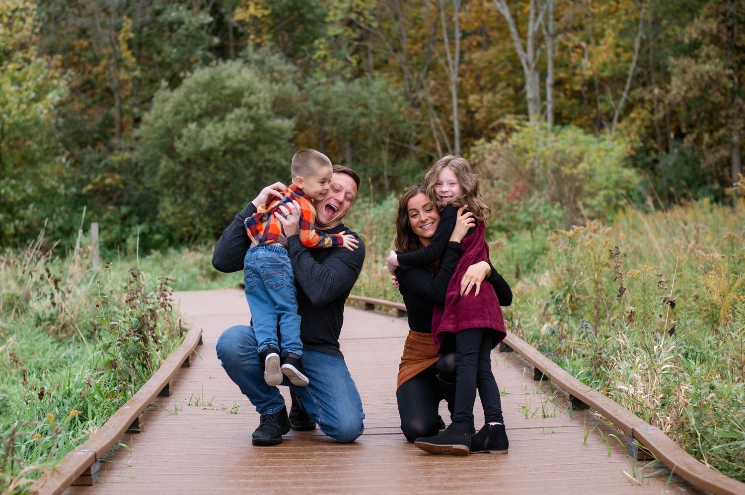 A couple sits together on a wooden boardwalk surrounded by autumn foliage in a natural outdoor setting.